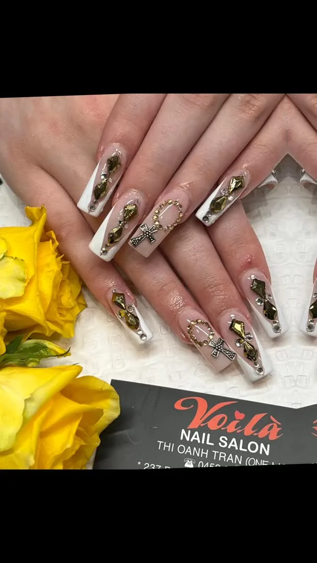 White-and-gold bejeweled long coffin nails with cross charms beside yellow roses